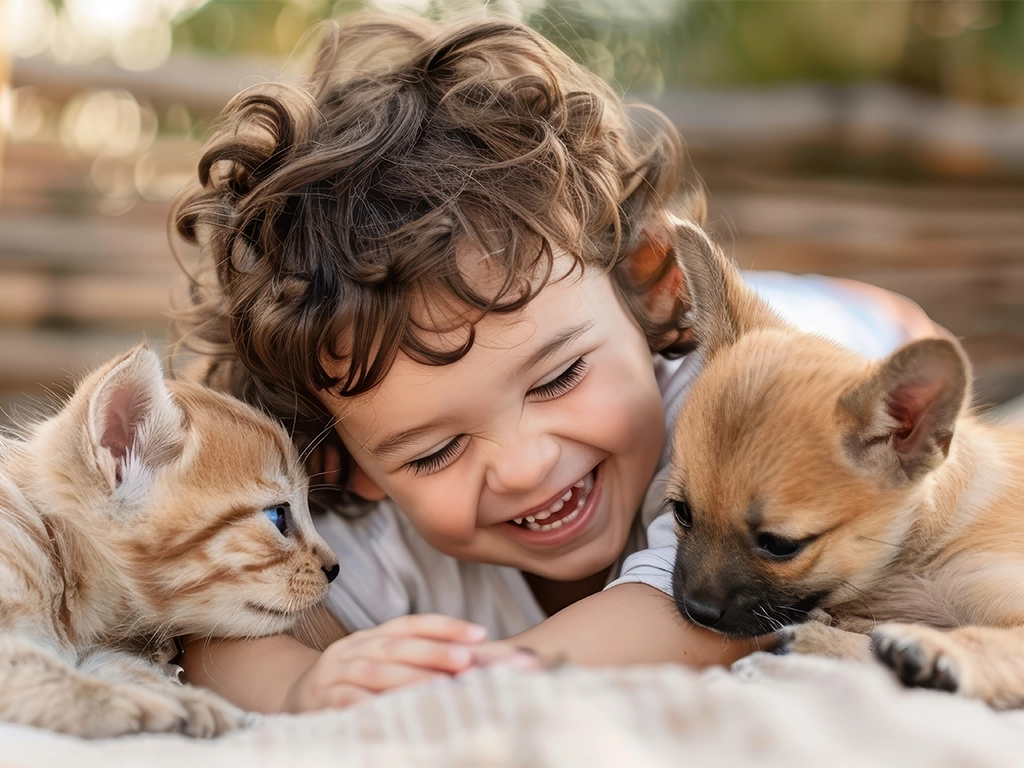 child lies on green grass, smiling while cuddling a kitten and a puppy